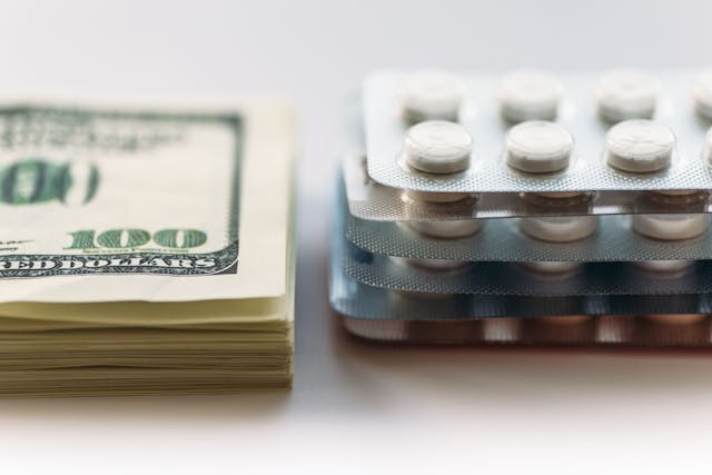 Image credit: DedMityay | stock.adobe.com. Bundle of money and pack of medication tablets or drug pills, close-up. Expensive health care concept